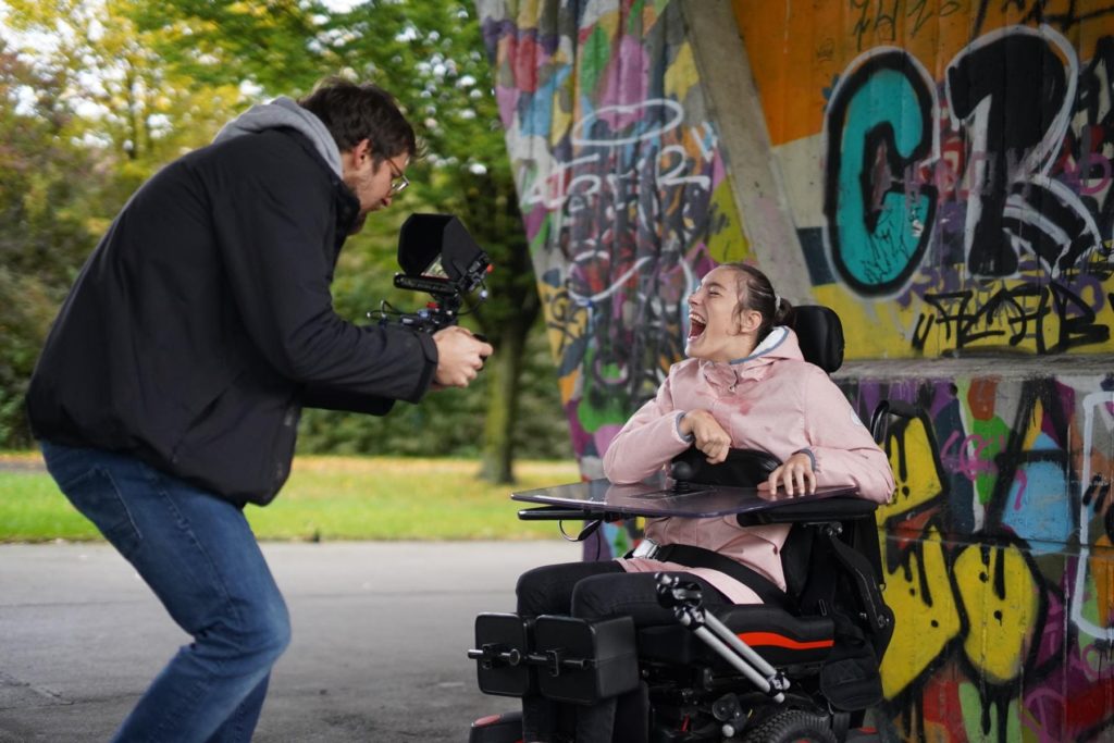 Eine Frau in einem speziellen Rollstuhl lacht und wird von einem Mann mit Kamera gefilmt