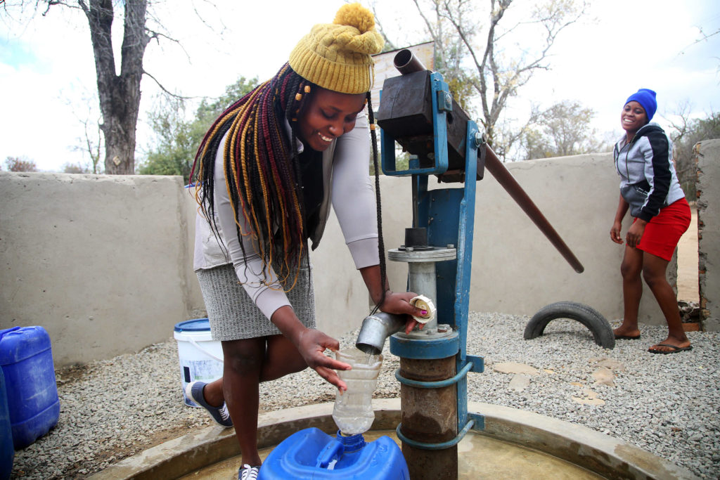 Frau mit Locks füllt Wasser aus Brunnen in eine Flasche, eine andere schaut ihr lächelnd zu. Foto: share