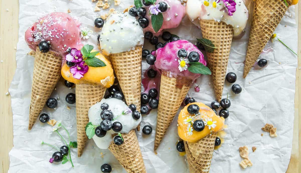 Mehrere Waffeltüten mit unterschiedlichen Eissorten liegen auf einem Tisch. Auf ihnen sind Blaubeeren und Glitzer verstreut.