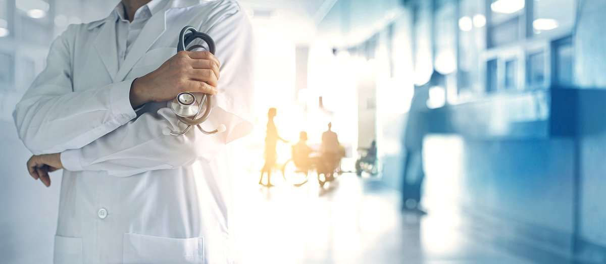 Vordergrund: Person in weißem Arztkittel hält ein Stethoskop. Im Hintergrund sind unscharf Patient:innen zu erkennen, die Multiple Sklerose haben könnten.