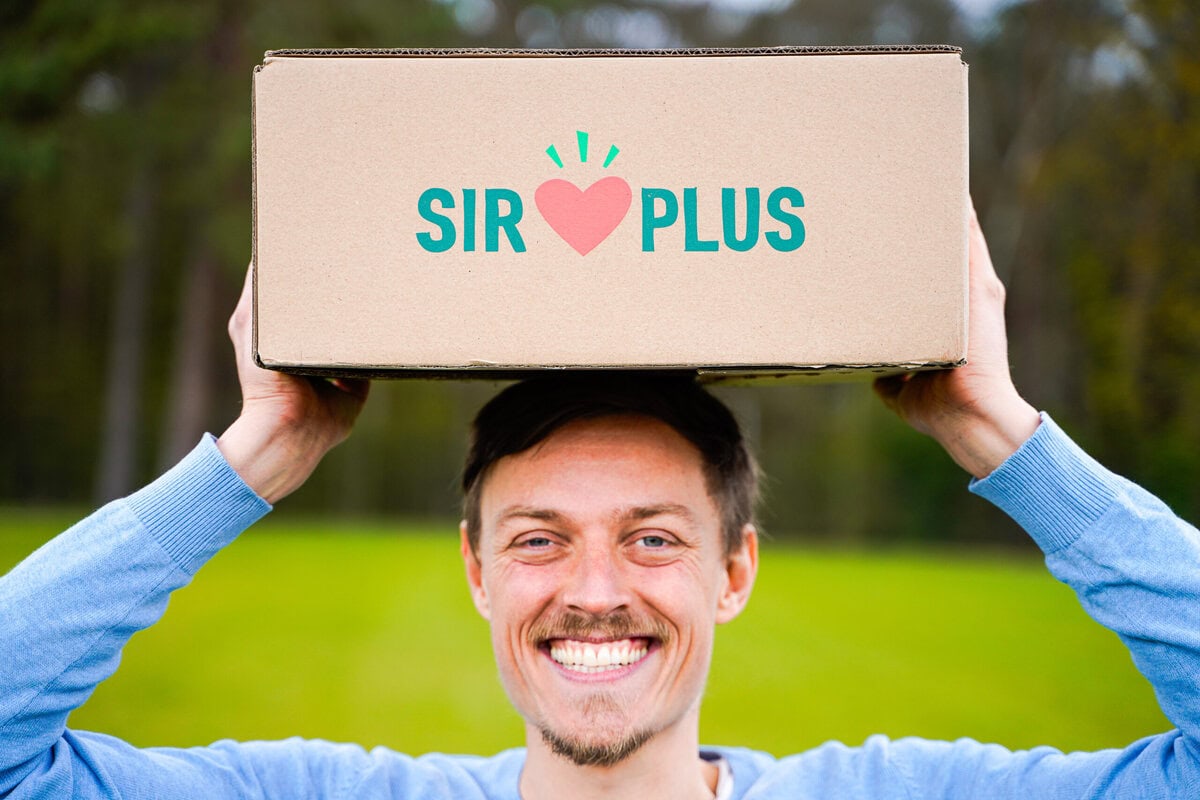 Der SIRPLUS-Mitgründer Raphael Fellmer hält eine Box mit geretteten Lebensmitteln hoch und strahlt in die Kamera. Foto von Hassaan Hakim
