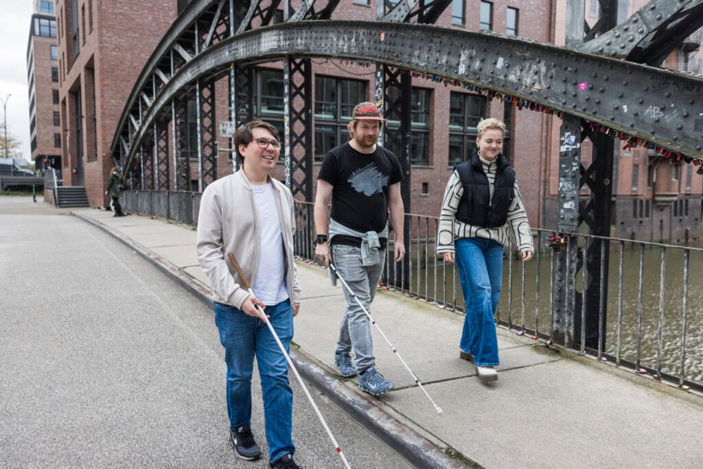 Menschen mit Sehbehinderung erkunden die Stadt an einem verregneten Tag, begleitet von einer Begleitperson im urbanen Umfeld.