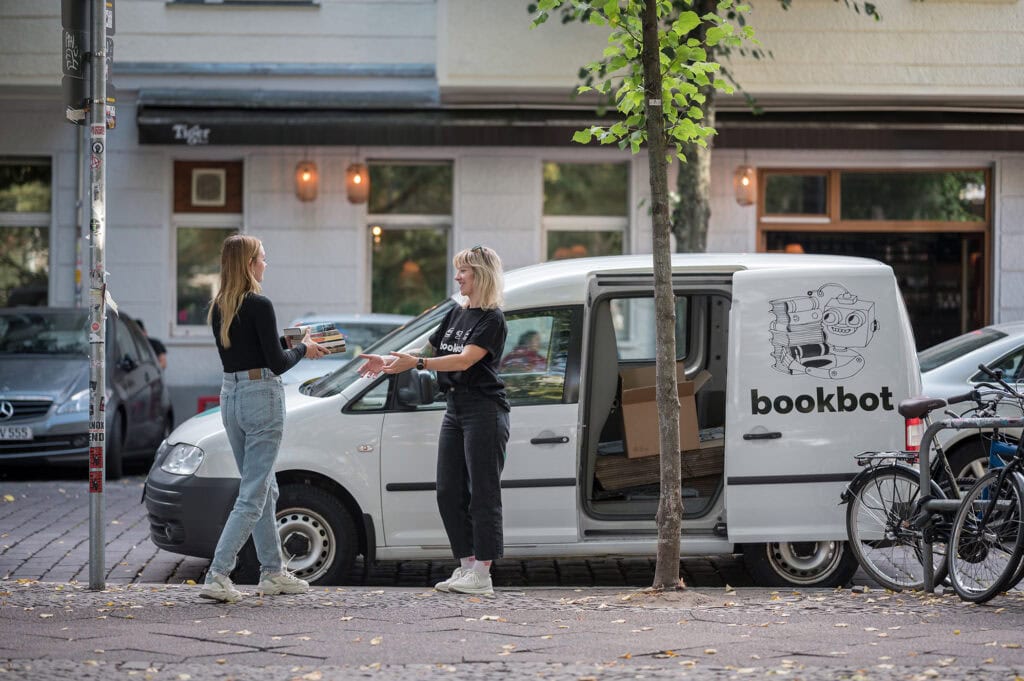 Re-Commerce im Buchhandel mit Bookbot. Eine Frau reicht einer anderen Bücher vor einem Bookbot-Lieferfahrzeug.
