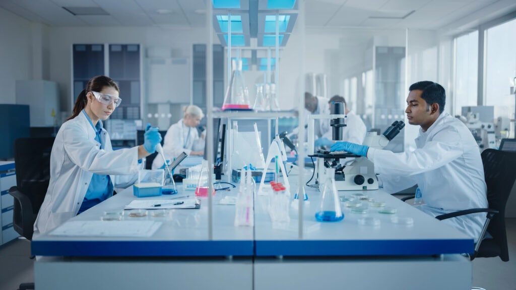 Long COVID und ME/CFS sollen besser erforscht werden. Bild von Menschen im Labor. Credits: https://depositphotos.com/de/photo/modern-medical-research-laboratory-diverse-team-of-scientists-working-with-pipette-analysing-biochemicals-samples-talking-453095236.html